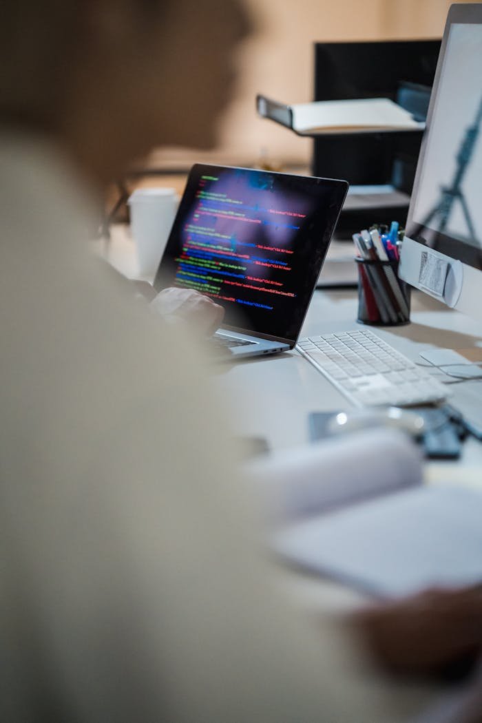 Blurred figure at a desk with a laptop displaying colorful code, indicating a programming environment.